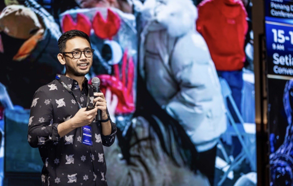 Abi Mangku with a speaker badge on stage at PDX2026, mid-talk