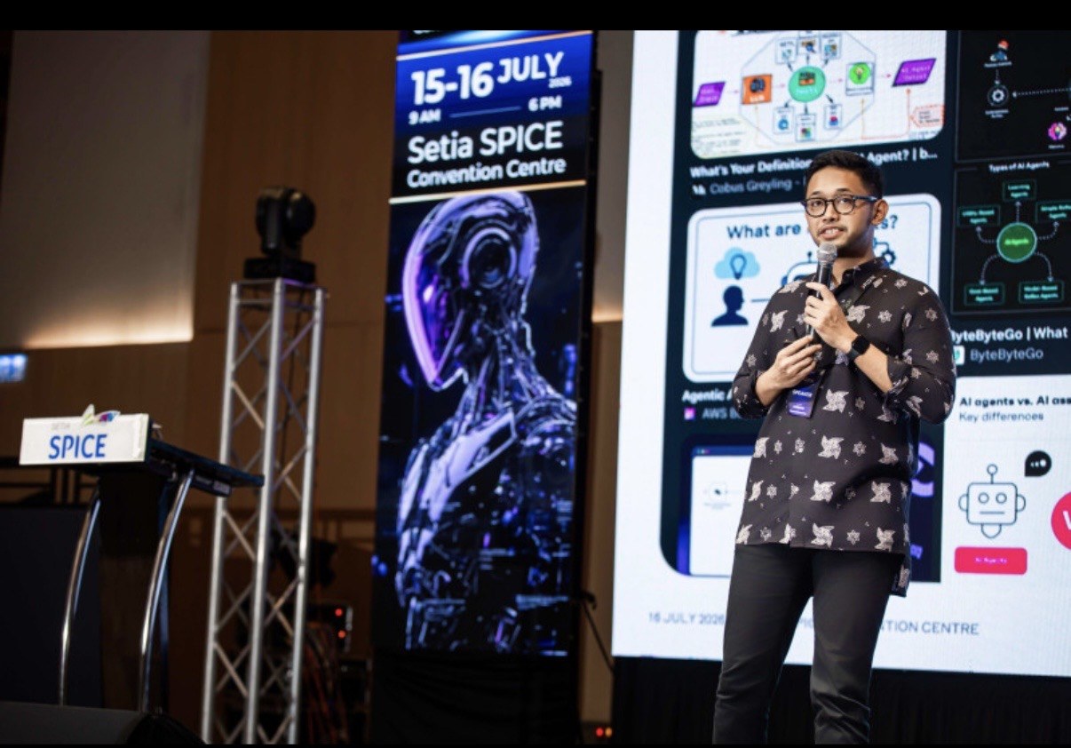 Abi Mangku speaking at PDX2026 beside the Setia SPICE Convention Centre event banner